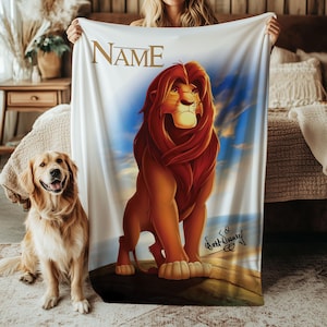 Puede incluir: Una manta blanca con una ilustración detallada de un león con melena roja y un fondo de cielo azul. La palabra "NAME" está impresa en oro en la parte superior. Un perro golden retriever está sentado en el lado izquierdo de la manta.