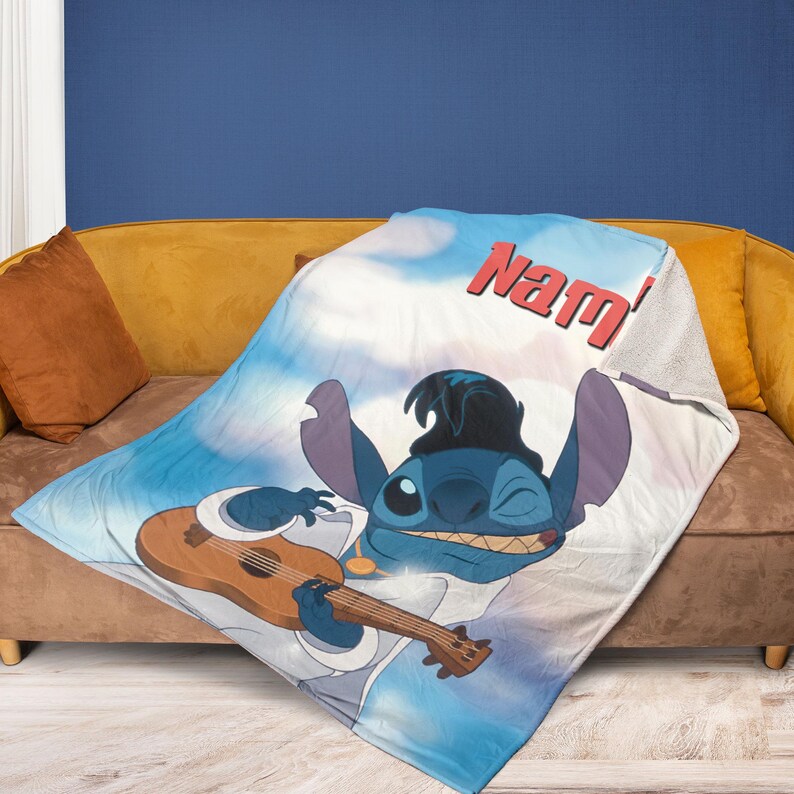 May include: A blue and white blanket featuring a cartoon character playing a ukulele. The character is blue with large ears and is wearing a hat and a white shirt. The blanket has the word "Nam'" in red.