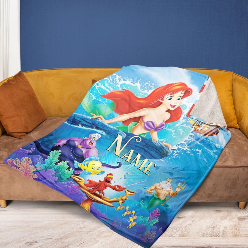 May include: A blue blanket featuring Ariel from The Little Mermaid, with other characters like Ursula, Flounder, and Sebastian. The blanket has the word "NAME" in gold lettering. The blanket is on a brown sofa.
