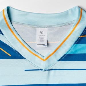 Puede incluir: Una camisa de rayas azules y azul claro con cuello en V y ribete naranja. La camisa tiene una etiqueta de talla que dice "M" y "GarmentOye".