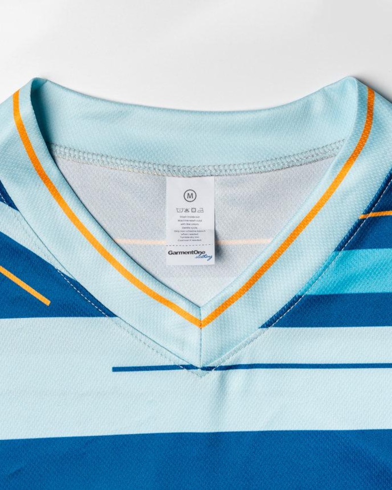 Puede incluir: Una camisa de rayas azules y azul claro con cuello en V y ribete naranja. La camisa tiene una etiqueta de talla que dice "M" y "GarmentOye".