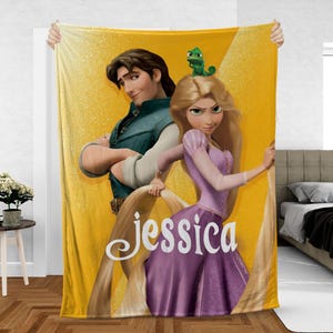 May include: A yellow fleece blanket featuring a cartoon illustration of a princess and a prince. The princess has long blonde hair and is wearing a purple dress. The prince is wearing a green vest. The name "Jessica" is written on the blanket.