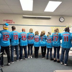 May include: A group of teachers wearing blue and red baseball jerseys with numbers on the back. The jerseys have the names "McCully", "Reed", "Maidrama", "Swisher", "Gibbons", "Tandolph", "Streby", and "Horning" on the back. The teachers are standing in a line in front of a white board with the words "Learning Targets" written on it.