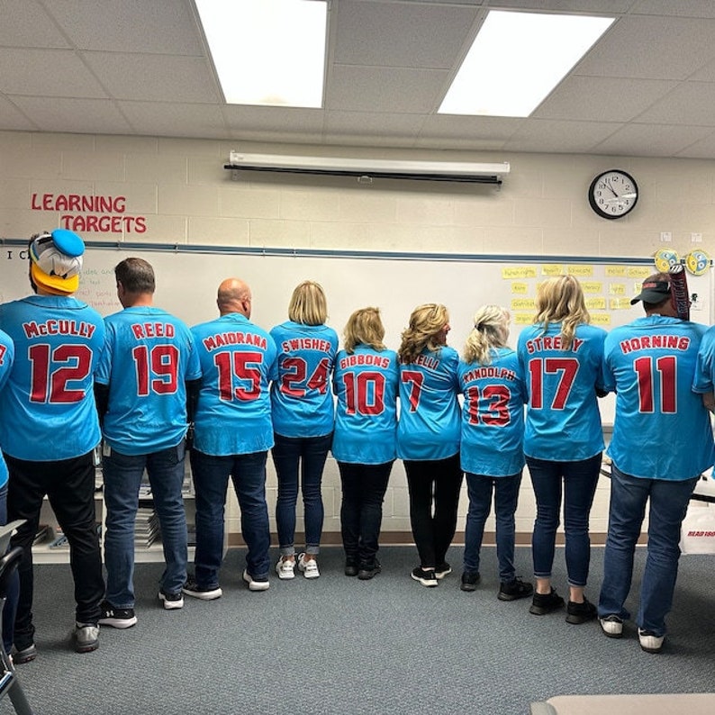 May include: A group of teachers wearing blue and red baseball jerseys with numbers on the back. The jerseys have the names "McCully", "Reed", "Maidrama", "Swisher", "Gibbons", "Tandolph", "Streby", and "Horning" on the back. The teachers are standing in a line in front of a white board with the words "Learning Targets" written on it.
