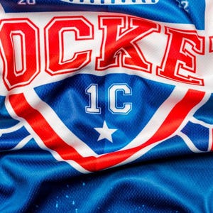 Puede incluir: Una camiseta de hockey blanca, roja y azul con la palabra "ROCKET" en letras rojas y blancas y el n&uacute;mero "1C" en letras blancas. La camiseta tiene una estrella en el centro del logotipo.