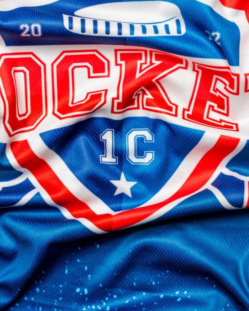 Puede incluir: Una camiseta de hockey blanca, roja y azul con la palabra "ROCKET" en letras rojas y blancas y el n&uacute;mero "1C" en letras blancas. La camiseta tiene una estrella en el centro del logotipo.