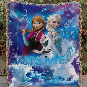May include: A woven throw blanket featuring characters from the animated film "Frozen." The design includes Anna, Elsa, Olaf, and ice-themed imagery. The word "NAME" is displayed in blue. The blanket has a fringed edge.