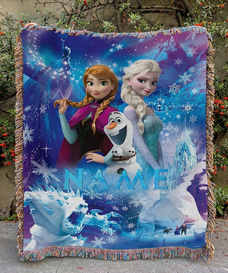 May include: A woven throw blanket featuring characters from the animated film "Frozen." The design includes Anna, Elsa, Olaf, and ice-themed imagery. The word "NAME" is displayed in blue. The blanket has a fringed edge.