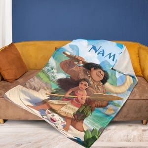 May include: A printed blanket featuring characters from the animated film "Moana." The design includes Moana, Maui, and Pua the pig, set against a tropical island backdrop with the word "Nam" visible.