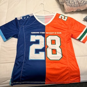 May include: A blue and orange football jersey with the number 28 on the front. The jersey is split down the middle, with the left side being blue and the right side being orange. The jersey has the words "Tennessee Titans" and "University of Miami" printed on the front.