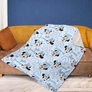 May include: A light blue blanket featuring a pattern of Minnie Mouse, bows, rainbows, and clouds. The name "Jenny" is written across the blanket. The blanket is draped over a brown couch with a yellow cushion.