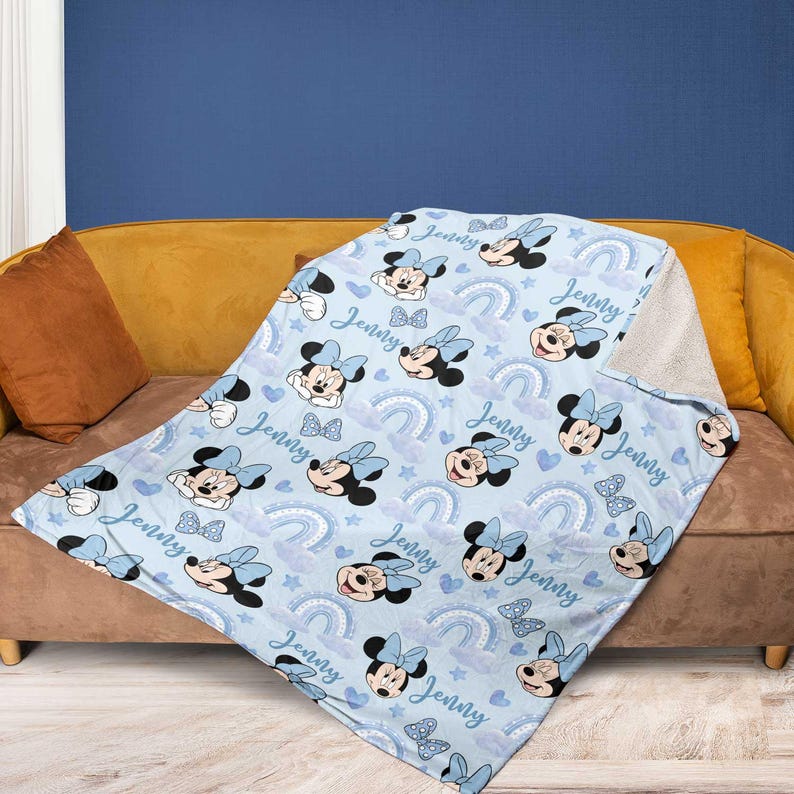 May include: A light blue blanket featuring a pattern of Minnie Mouse, bows, rainbows, and clouds. The name "Jenny" is written across the blanket. The blanket is draped over a brown couch with a yellow cushion.