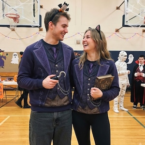 May include: Two matching dark purple sweatshirts featuring a printed design of a formal vest, blazer, and tie. The sweatshirts include a silver chain graphic, with one person holding a plastic hook and the other holding a vintage-style book.