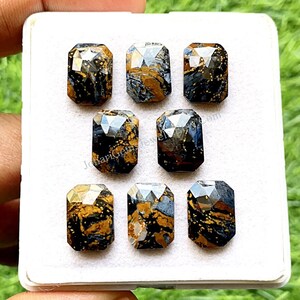 Puede incluir: Ocho cabujones de piedra preciosa rectangulares y facetados en una caja blanca. Las piedras preciosas son una mezcla de colores negro, dorado y azul. Las piedras preciosas están dispuestas en dos filas de cuatro.