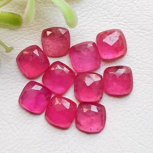 May include: Ten pink ruby gemstone cabochons, each with a square shape and faceted edges. The stones are arranged in a loose cluster on a white surface.