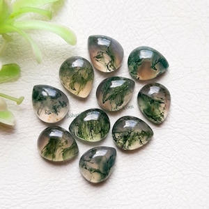 Puede incluir: Colección de nueve piedras preciosas de ágata musgosa pulidas. Cada piedra, en forma de lágrima, muestra un aspecto translúcido con inclusiones verdes, blancas y marrones que se asemejan al musgo. Las piedras están dispuestas sobre una superficie blanca.