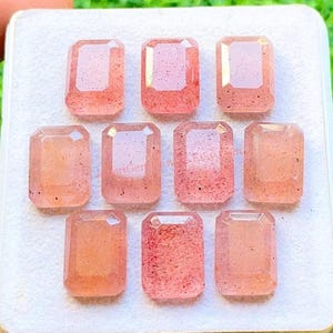 Puede incluir: Nueve gemas rosas claras, de corte rectangular, con motas internas, están dispuestas sobre una superficie blanca. Las gemas tienen un aspecto pulido y transparente, probablemente para la fabricación de joyas o coleccionismo.