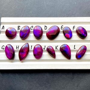 Puede incluir: Una colección de doce piedras preciosas pulidas, cada una con una forma única y una vibrante paleta de colores púrpura, magenta y rojo intenso. Las piedras están dispuestas en una bandeja blanca, mostrando sus características y variaciones individuales.