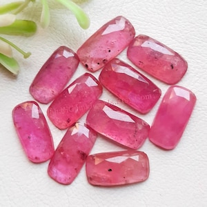 May include: A collection of ten rectangular, faceted pink gemstones with visible dark inclusions. Each stone features a polished surface with geometric cuts that reflect light, arranged in a loose cluster on a textured white surface.