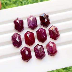 Könnte beinhalten: Zehn facettierte Rubingesteine in verschiedenen Rottönen, angeordnet auf einer weißen Oberfläche.