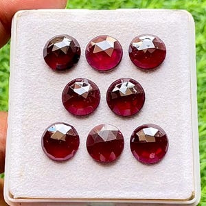May include: Seven round, faceted, deep red gemstones arranged in a grid pattern on a white, textured display pad. Each stone features a brilliant cut with multiple triangular facets that reflect light, creating a sparkling, dark crimson appearance.