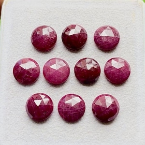 Könnte beinhalten: Neun runde, facettierte Rubine in einem Raster angeordnet. Jeder Edelstein hat eine tiefrote Farbe mit einer Rosenschliff-Oberfläche und zeigt mehrere dreieckige Facetten, die das Licht auf den runden, polierten Flächen der Steine reflektieren.