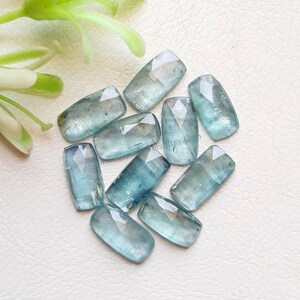 May include: A group of ten light blue aquamarine gemstones, cut into rectangular shapes with faceted edges. The stones are arranged on a white surface.