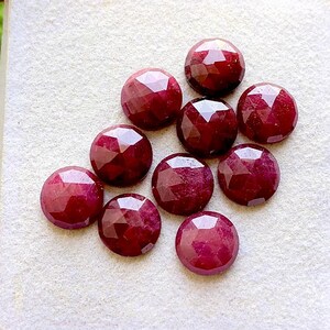 Puede incluir: Un conjunto de nueve rubíes redondos y facetados de color rojo intenso. Cada piedra preciosa presenta una superficie geométrica de múltiples facetas que refleja la luz, dispuestas en un grupo sobre una superficie texturizada de color claro.