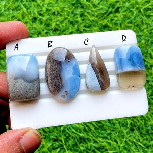 Puede incluir: Cuatro piedras preciosas pulidas, cada una con patrones y colores únicos, presentadas en un soporte blanco. Las piedras, etiquetadas A, B, C y D, muestran tonos de azul, marrón y gris. El fondo es verde.