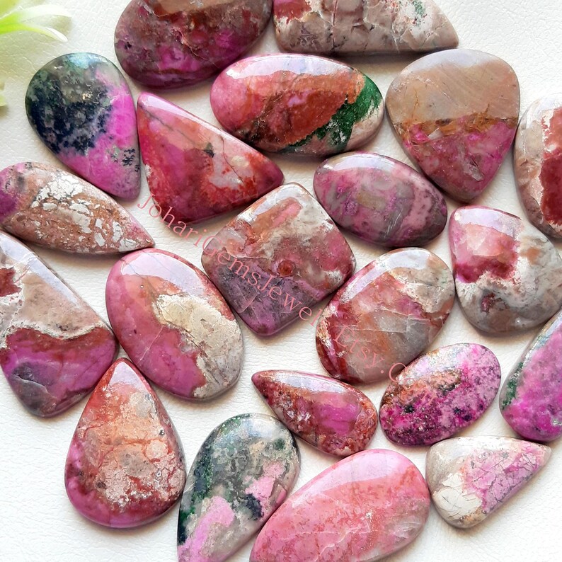 May include: A collection of 15 pink, red, and white cabochon gemstones with green and brown inclusions. The stones are various shapes and sizes, including teardrop, oval, and square.