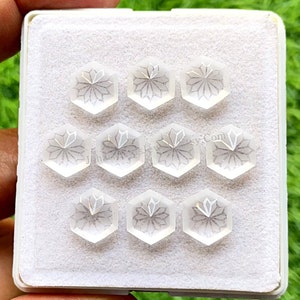 May include: Ten clear, hexagonal, faceted gemstones with a carved flower design. The gemstones are arranged in a grid pattern in a white box.