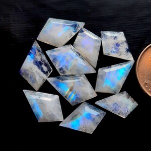 Puede incluir: Un conjunto de ocho piedras de luna facetadas, cada una cortada en una forma única de cometa o diamante. Estas piedras translúcidas exhiben una distintiva adularescencia azul, reflejando la luz con destellos iridiscentes. Una moneda de cobre es visible para la escala.