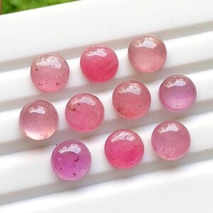 May include: Nine round, polished pink gemstones arranged in a grid. Each stone features a smooth, domed cabochon surface with varying shades of translucent pink, ranging from pale rose to deep magenta, showing natural internal inclusions and reflections.