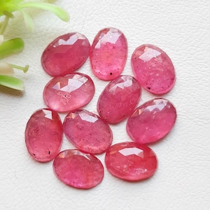 Puede incluir: Un conjunto de nueve piedras preciosas rosas de forma ovalada, facetadas y con inclusiones oscuras visibles. Las piedras están dispuestas en un grupo, cada una mostrando una superficie pulida con facetas geométricas que reflejan la luz.