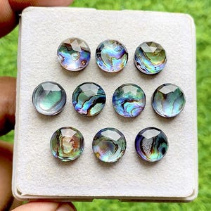 Peut inclure: Neuf cabochons ronds en coquille d'ormeau à facettes disposés en grille sur un support en mousse blanche. Chaque cabochon présente un motif irisé unique avec des tourbillons de bleu, vert, violet et or, reflétant la lumière sur la surface.