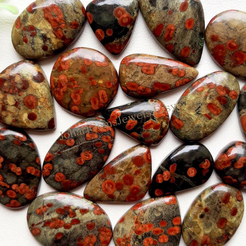 May include: A collection of polished gemstones with a speckled pattern. The stones vary in shape, including oval, teardrop, and round. The colors are primarily shades of brown, gray, and black, with red spots throughout. The gemstones are likely for jewelry making.