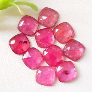 May include: A collection of ten faceted, cushion-cut pink gemstones arranged in a cluster. Each stone features a square shape with rounded corners and a polished surface, displaying varying shades of vibrant pink with subtle internal inclusions.