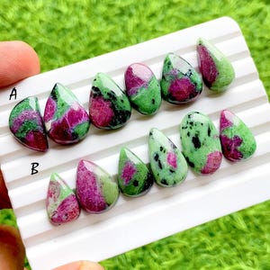 Peut inclure: Une collection de pierres précieuses en rubis zoïsite polies, disposées en deux rangées sur un plateau blanc. Les pierres présentent un mélange d'inclusions de rubis magenta vif et de zoïsite verte avec des taches noires, en forme de goutte.