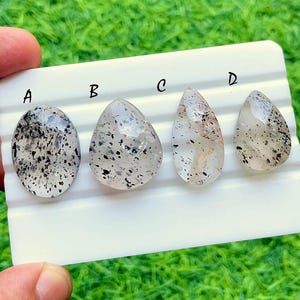 May include: Four polished, translucent gemstones labeled A, B, C, and D, arranged on a white display stand. Gemstone A is oval, while B, C, and D are teardrop-shaped. Each stone features internal black speckles and inclusions throughout.