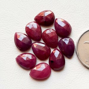 Pode incluir: Dez pedras preciosas de rubi facetadas em forma de pêra em vários tons de vermelho. As pedras preciosas estão dispostas em um cacho sobre uma superfície branca.