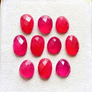 May include: Ten faceted, oval-shaped, vibrant pinkish-red gemstones arranged in three rows. Each stone features a polished surface with geometric facets that reflect light, showcasing a consistent deep red color and a smooth, rounded edge profile.