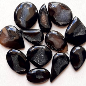 May include: A collection of 12 black agate cabochons with a druzy crystal pattern. The cabochons are various shapes and sizes, including oval, teardrop, and round.