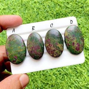 Può includere: Quattro pietre preziose ovali con un colore di base verde e inclusioni rosse e dorate. Le pietre preziose sono etichettate A, B, C e D e sono esposte su una superficie bianca. Le pietre sono tenute da una mano.
