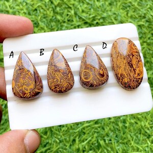 May include: Four polished gemstones with a teardrop shape, each displaying a unique pattern of brown and gold swirls. The gemstones are labeled A, B, C, and D, and are set against a white background.