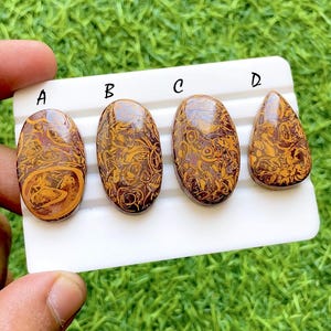 Puede incluir: Cuatro piedras preciosas pulidas con un patrón arremolinado marrón y dorado se muestran sobre una superficie blanca. Las piedras preciosas tienen forma ovalada y de lágrima. Las piedras preciosas están etiquetadas A, B, C y D.