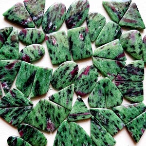 May include: A collection of polished zoisite gemstones with ruby inclusions. The stones are various shapes, including ovals, rectangles, and triangles. The dominant colors are green and red, with black speckling throughout.
