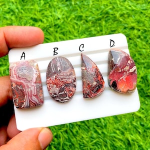 May include: Four polished gemstones with unique patterns and colors. The stones are labeled A, B, C, and D. The gemstones feature shades of red, brown, and cream, with intricate veining and patterns. The stones are displayed on a white surface against a green background.