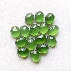 Op de afbeelding: Een verzameling van 14 groene, ovale edelstenen met gefacetteerde randen. De stenen liggen los op een wit oppervlak.