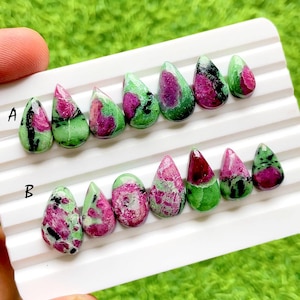 Peut inclure: Une collection de pierres précieuses en rubis zoïsite polies, disposées en deux rangées sur un plateau blanc. Les pierres présentent un mélange d'inclusions de rubis magenta vif et de zoïsite verte avec des taches minérales noires.