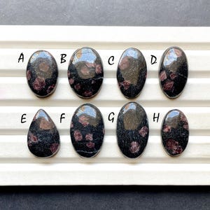 Peut inclure: Huit pierres précieuses polies à l'aspect sombre et moucheté. Les pierres sont de forme ovale et en goutte, avec un mélange de teintes sombres et brun rougeâtre. Chaque pierre est étiquetée d'une lettre de A à H.